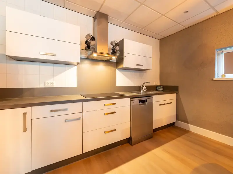 Witte keuken met kookplaat, stalen afzuigkap met mokken ernaast, spoelbak en vaatwasser.