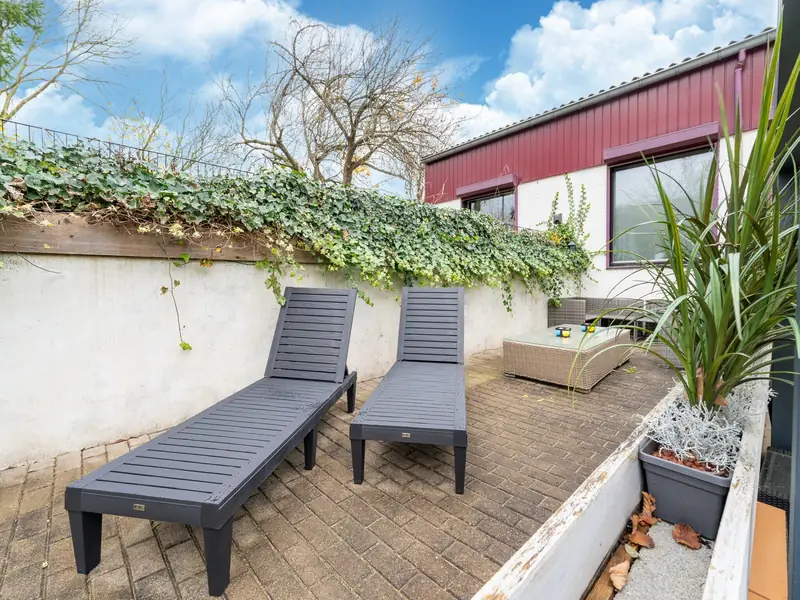 Terras met twee grijze ligbedden op klinkers, een witte muur met klimop en blauwe lucht.