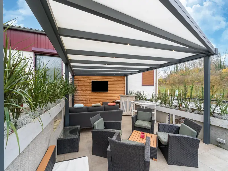Overdekte veranda met wicker loungestoelen, loungebank en tv, met bomen en gras op de achtergrond.