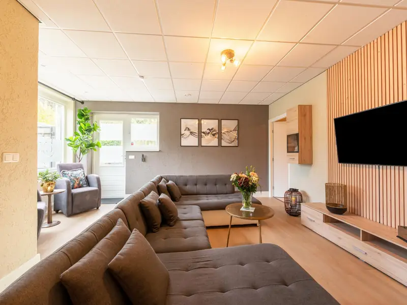 Woonkamer met grijze hoekbank, fauteuil bij het raam en tv aan de wand.
