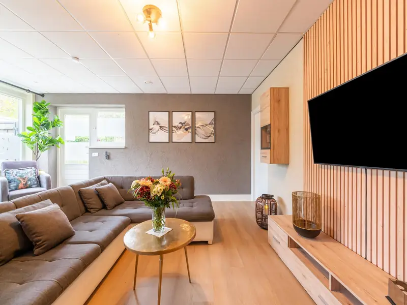 Woonkamer met grijze hoekbank, ronde goudkleurige salontafel en tv aan een houten latjeswand.