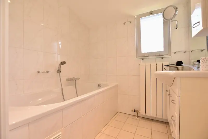 Witte badkamer met ligbad en handdouche, radiator, wastafelmeubel en een klein raam.