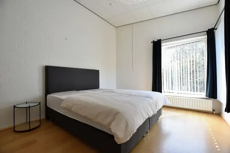 Slaapkamer met tweepersoonsbed, rond bijzettafeltje en een groot raam met lamellen.