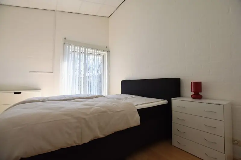 Slaapkamer met tweepersoonsbed, witte ladekast met rode lamp en een raam met lamellen.