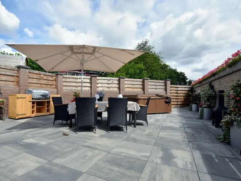 Groot betegeld terras met een tafel onder een parasol, een afgedekt bubbelbad en bloeiende planten.